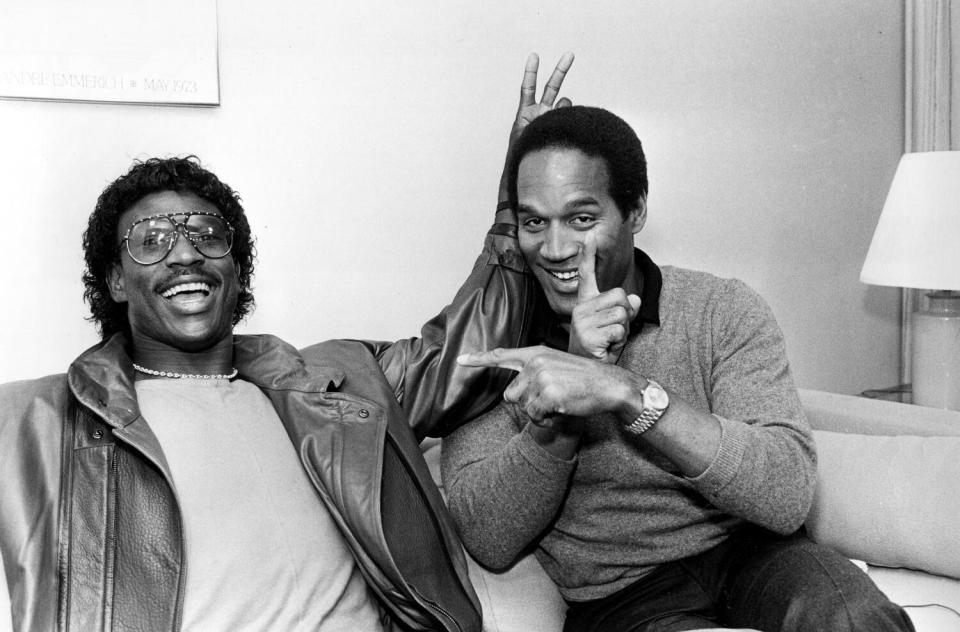 A black-and-white portrait of Eric Dickerson, left, and O.J. Simpson smiling on a hotel couch