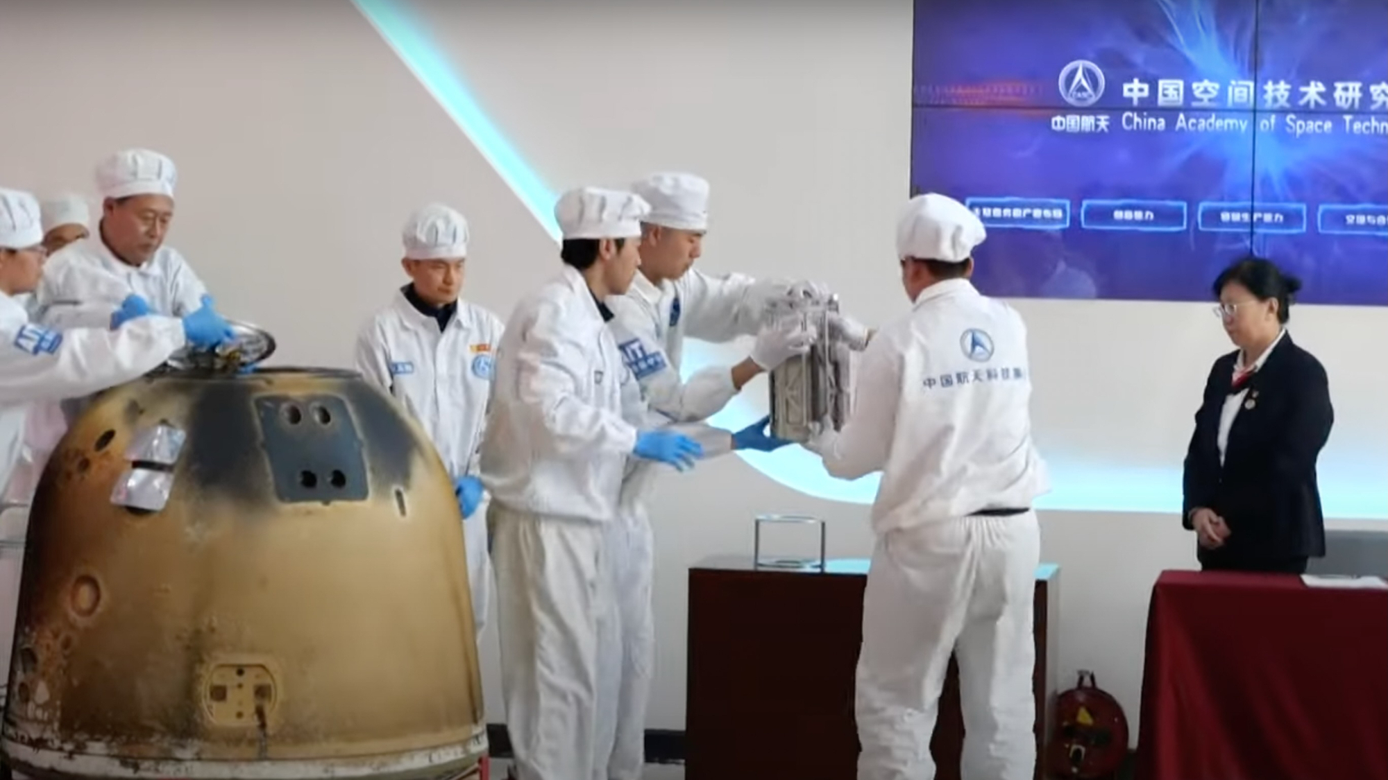 white-suited technicians remove a silver box from a gold spacecraft inside a white-walled room