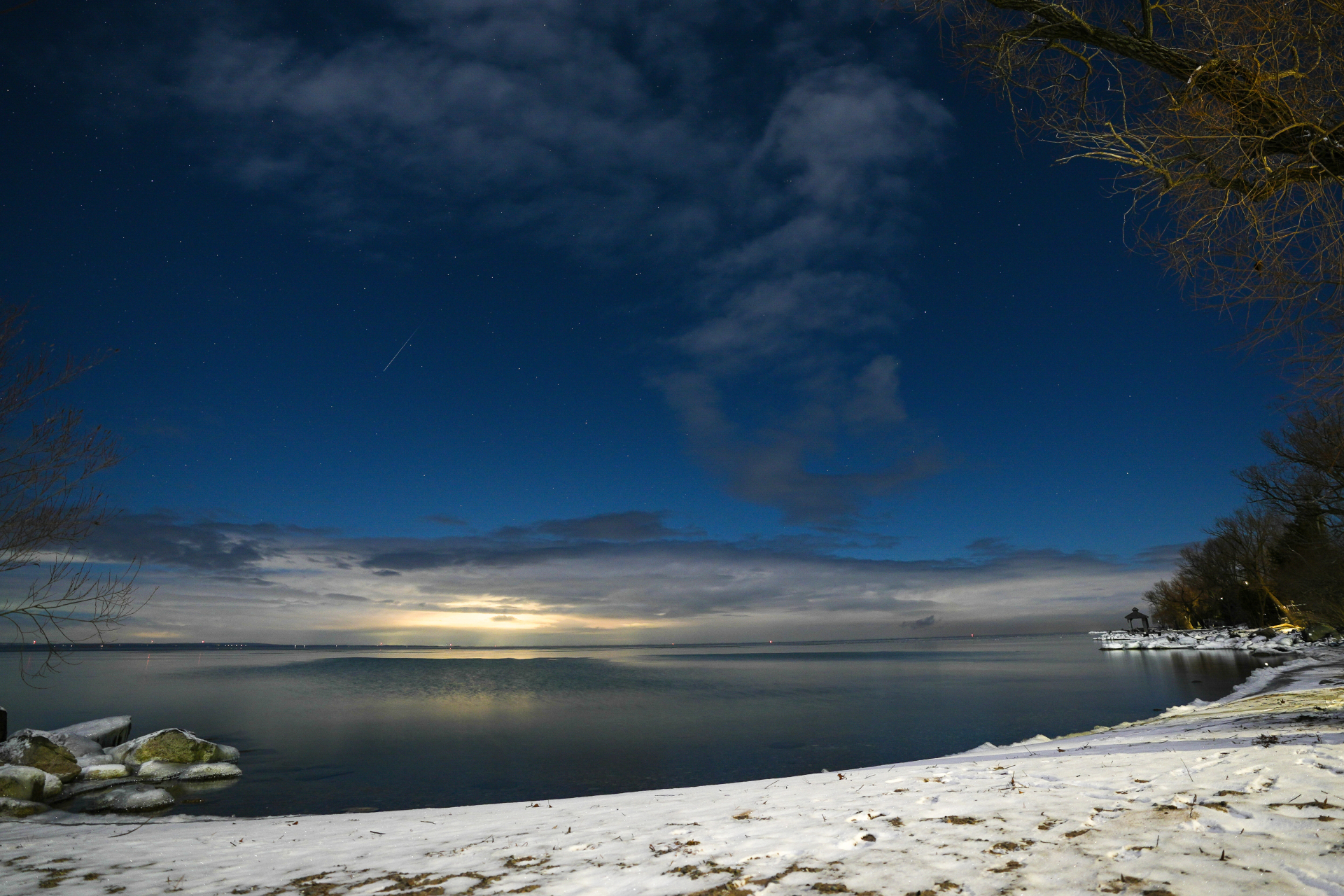 A meteor streaks across the night sky over Lake Simcoe, snow lays on the ground in the foreground and distant clouds line the sky.