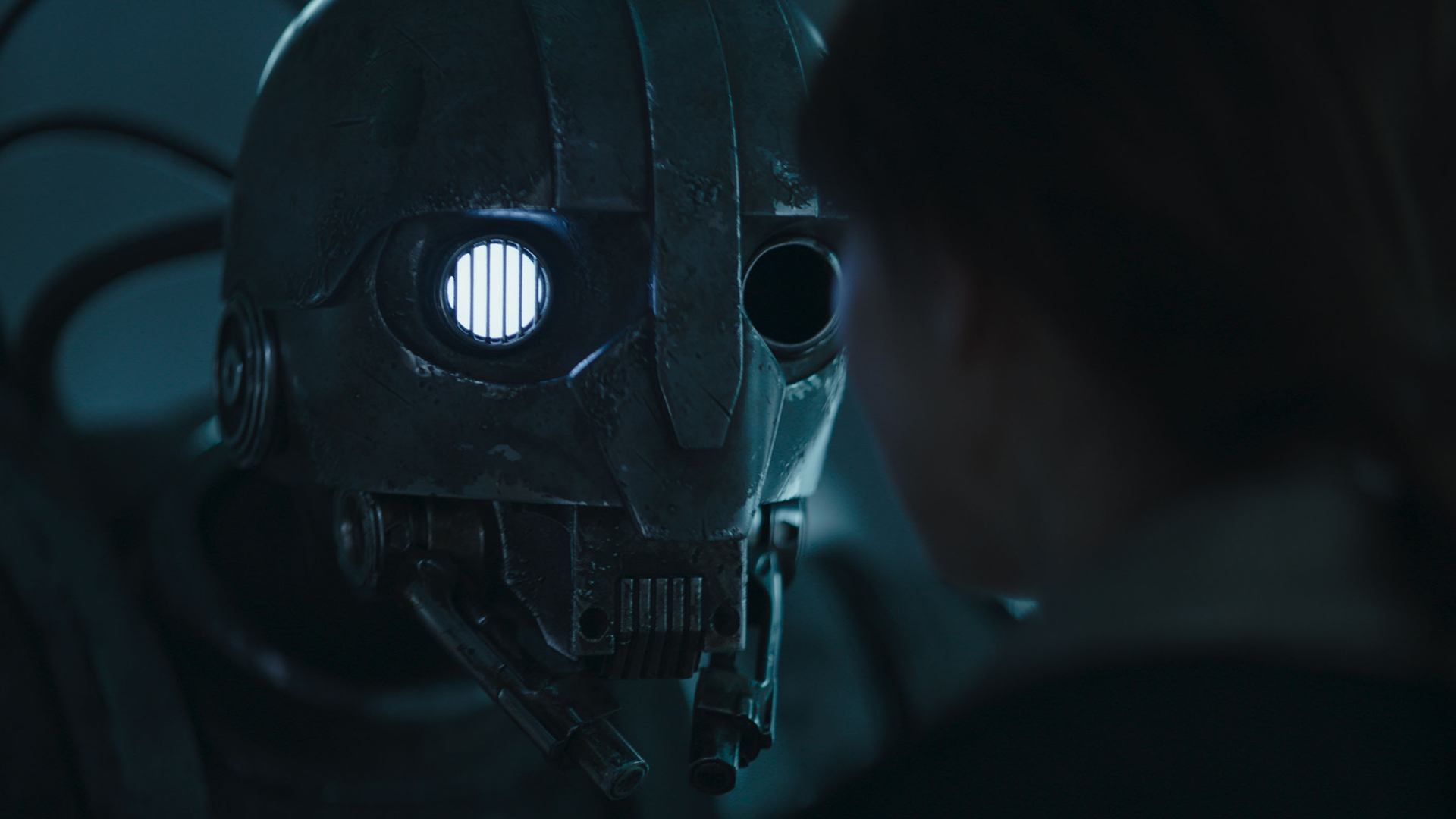 Still from Star Wars: Skeleton Crew TV series. Close up of SM-33, a humanoid droid with one eye lit up white and the other missing as he menacingly stares into Fern's (human girl) face.