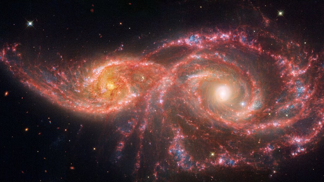 adjacent spiral galaxies with glowing arms of reds and pinks stare like a pair of eyes, their centers white hot yellow, from the black abyss of space.