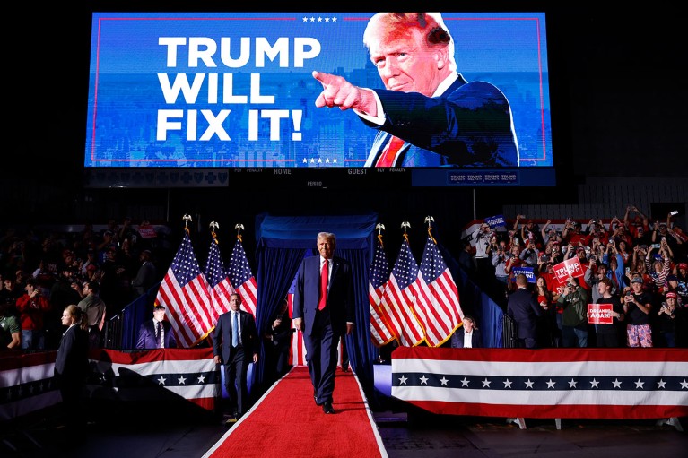 Beneath a large digital screen displaying his face, President-Elect Donald Trump walks out onto a stage.