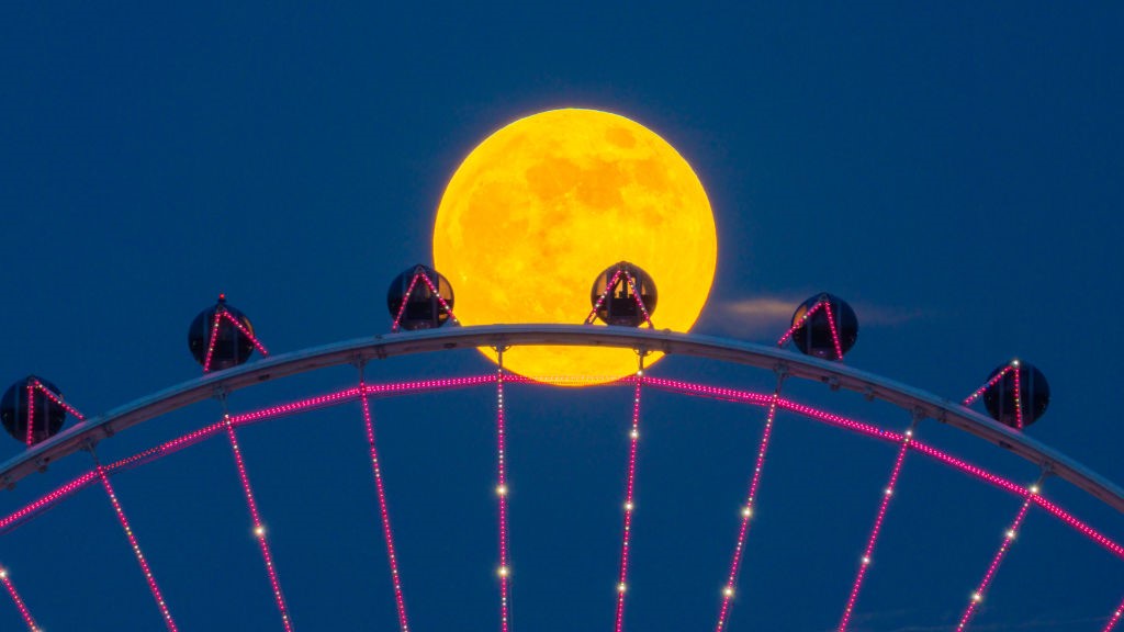 bright full moon behind a Ferris wheel with pink lights.