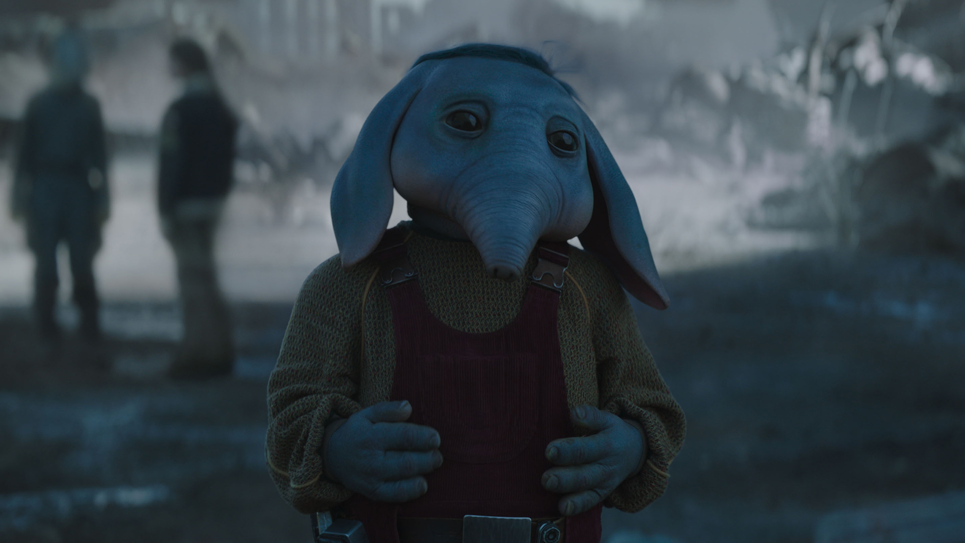 Still from Star Wars: Skeleton Crew TV series. Close up of Neel (blue elephant-like alien boy). He is wearing a brownish-yellow long-sleeved sweater cover by red overalls.