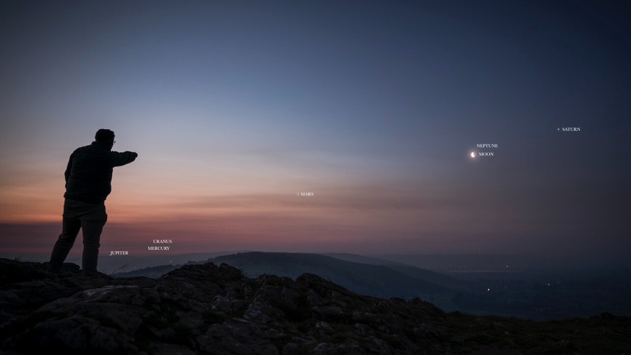 a person stands with their back to the viewer, pointing at several bright dots in the early morning sky