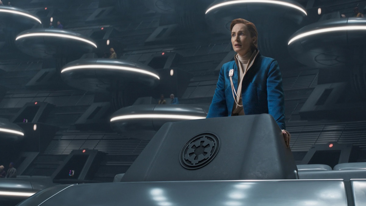 A woman in a blue outfit delivers a speech in a sci-fi senate chamber