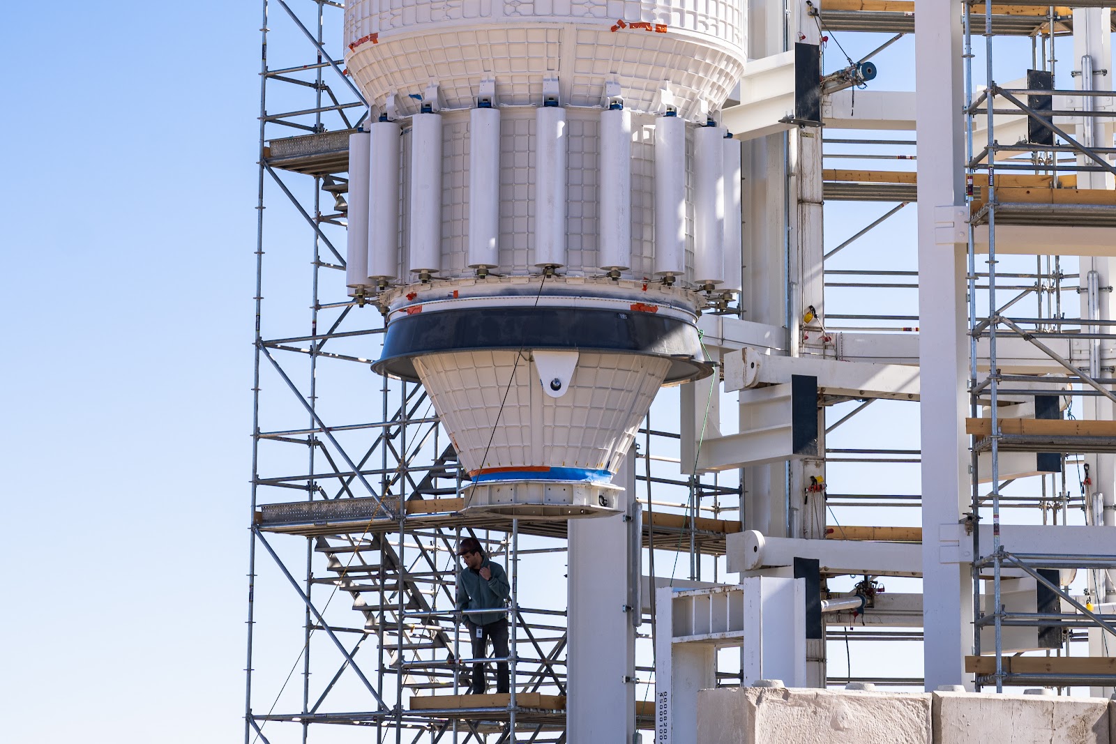 The conical bottom of a white space module is suspended in the desert against a bright blue sky, next to tall stacks of scaffolding.