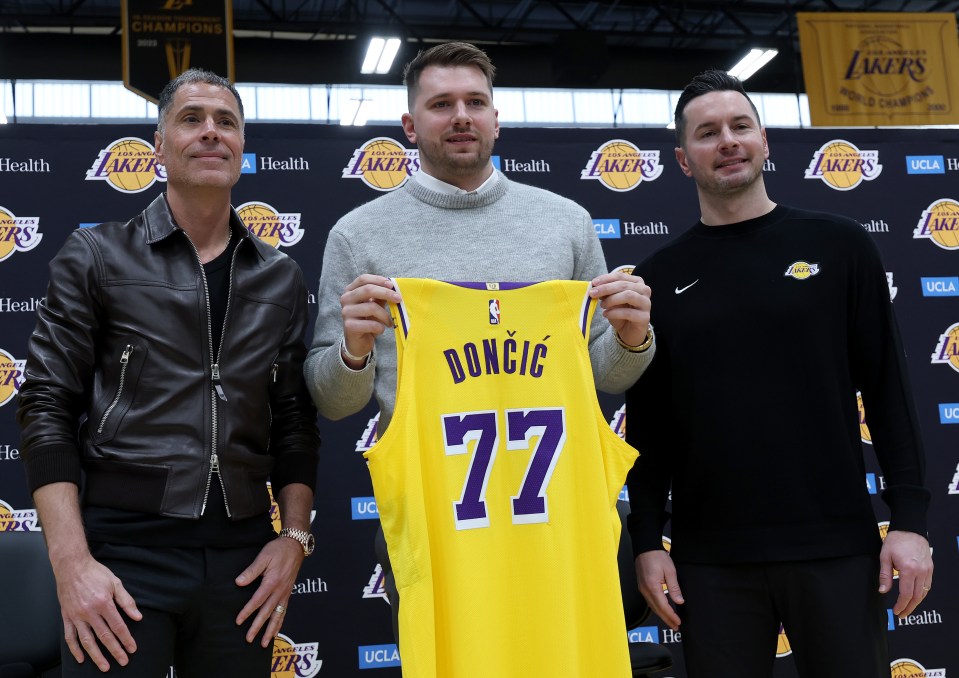 EL SEGUNDO, CALIFORNIA - FEBRUARY 04: Luka Doncic (C) of the Los Angeles Lakers holds his new jersey while standing alongside general manager Rob Pelinka (L) and head coach JJ Redick (R) during a press conference at UCLA Health Training Center on February 04, 2025 in El Segundo, California. (Photo by Harry How/Getty Images)