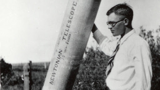 A black and white photo of a man with glasses next to a telescope that says 