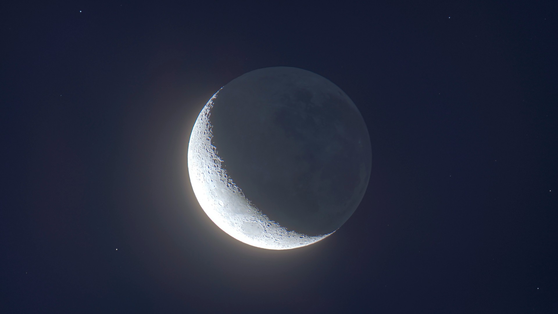 a grey orb, partially lit on its left side revealing craters and ridges. its right side is visible in a pale blue glow