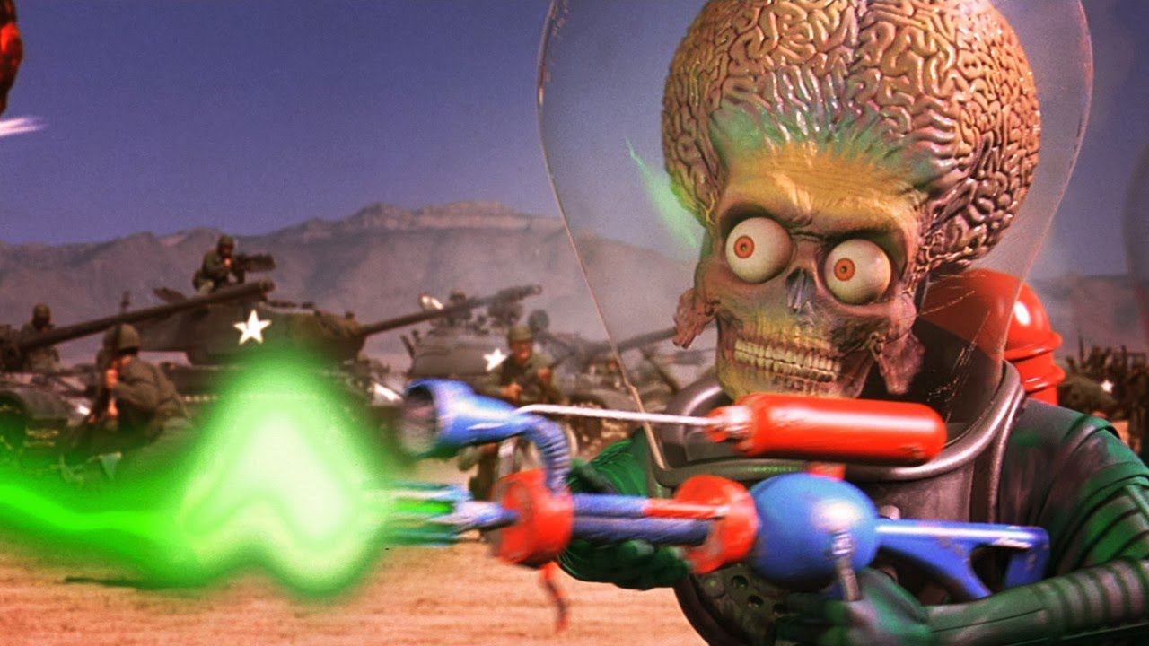 Screenshot from the movie Mars Attacks! (1996). An alien with a big bulging brain is firing a laser weapon. In the background there are several army tanks.