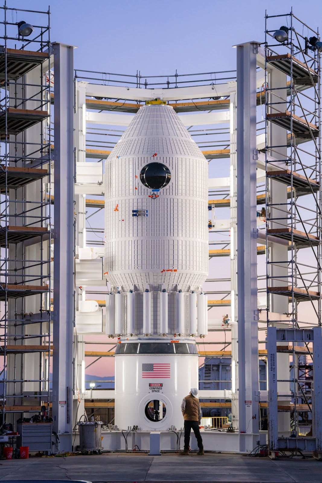 a white cylindrical vessel with conical top is mounted to stand on a test area, surrounded by railed scaffolding.