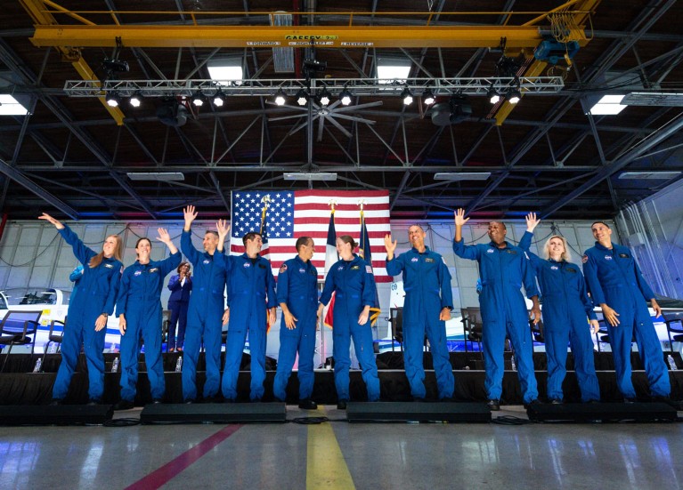 A wide-angle shot of ten NASA astronaut candidates standing in a line and waving, with the US flag in the background.