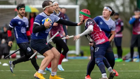 Giordano Di Pino-Forino A player cradles a ball while trying to guard it from an opponent. Their hand is outstretched and nearly touches the other player's shoulder. Three other players can be seen in the background coming to their aid.