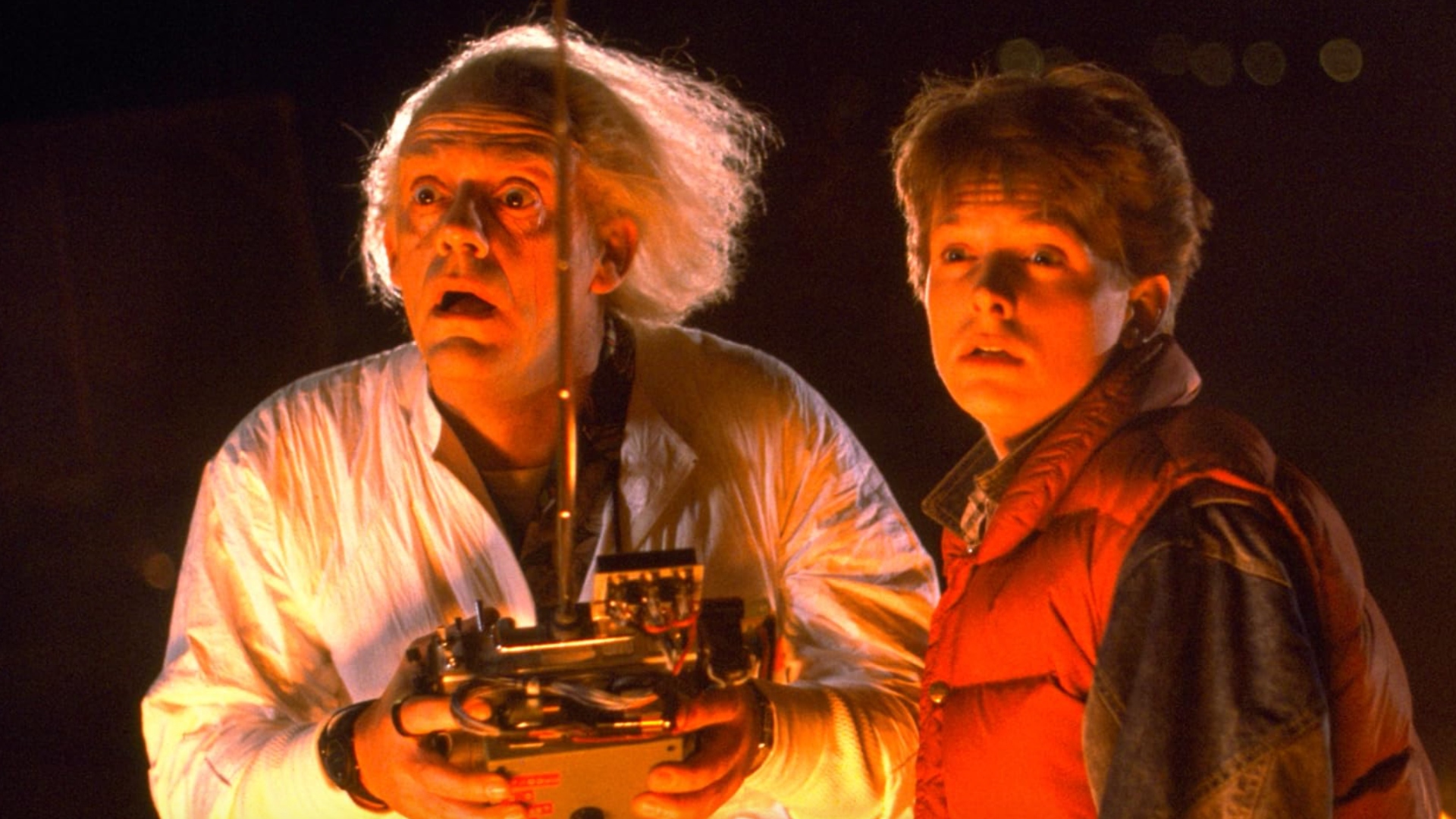 Screenshot from the movie Back to the Future. Doc Brown and Marty McFly are standing together in awe, basked in a glow of orange/red light. Doc Brown has long wild white hair and is wearing a white lab coat and holding a large remote controller in his hand.