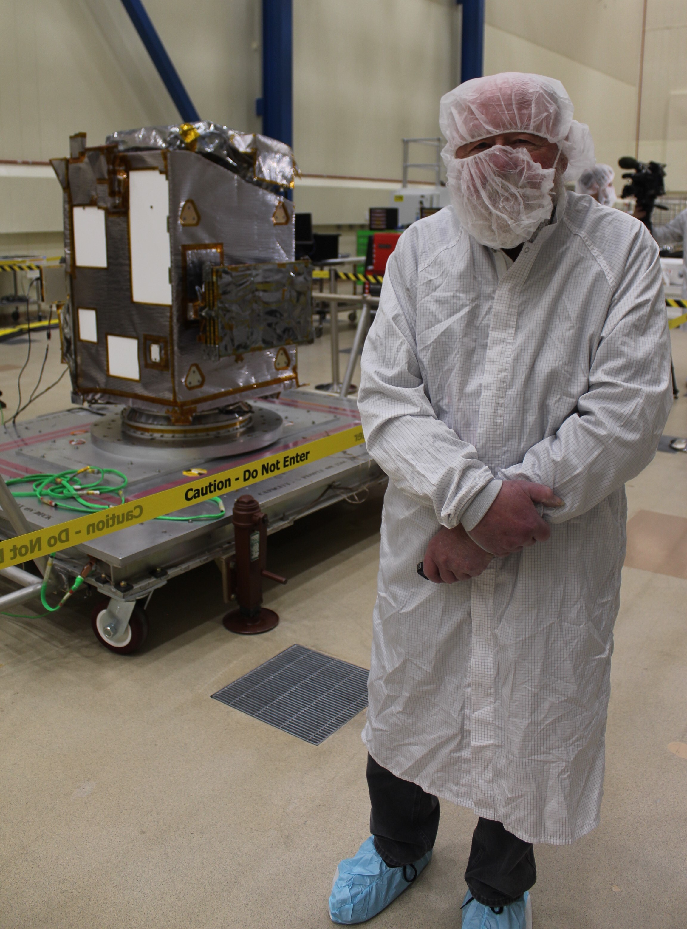 a bipedal organism stands adorned in a white coverall robe, with a hairnet worn on the top and front of their head. They stand next to a cube-shaped space probe sitting on metal slab with wheels.