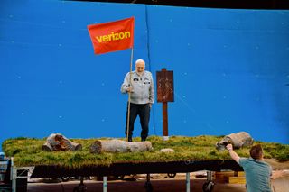 a white-haired man in a silver jacket and black pants holds a red and yellow flag backdropped by a blue screen on the set of a commercial.