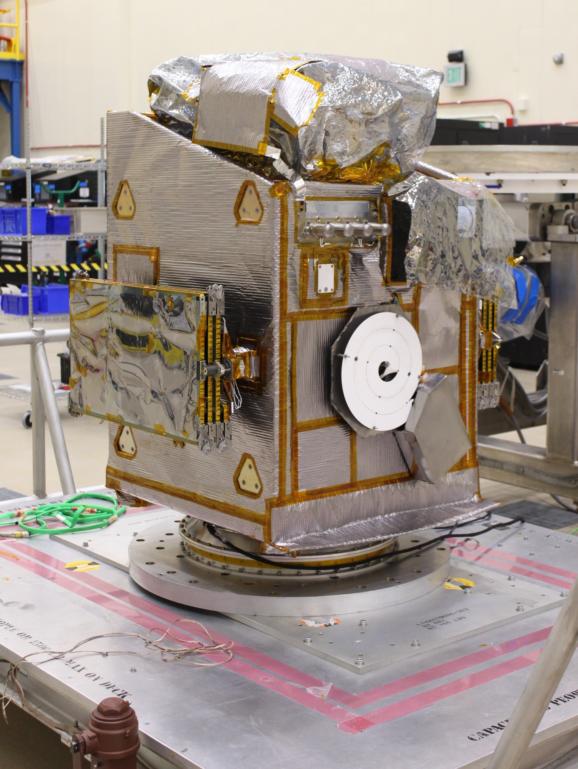 A big metal space probe shaped like an odd cube of sorts is wrapped in chrome foil, sitting on a metal slab in a clean room.